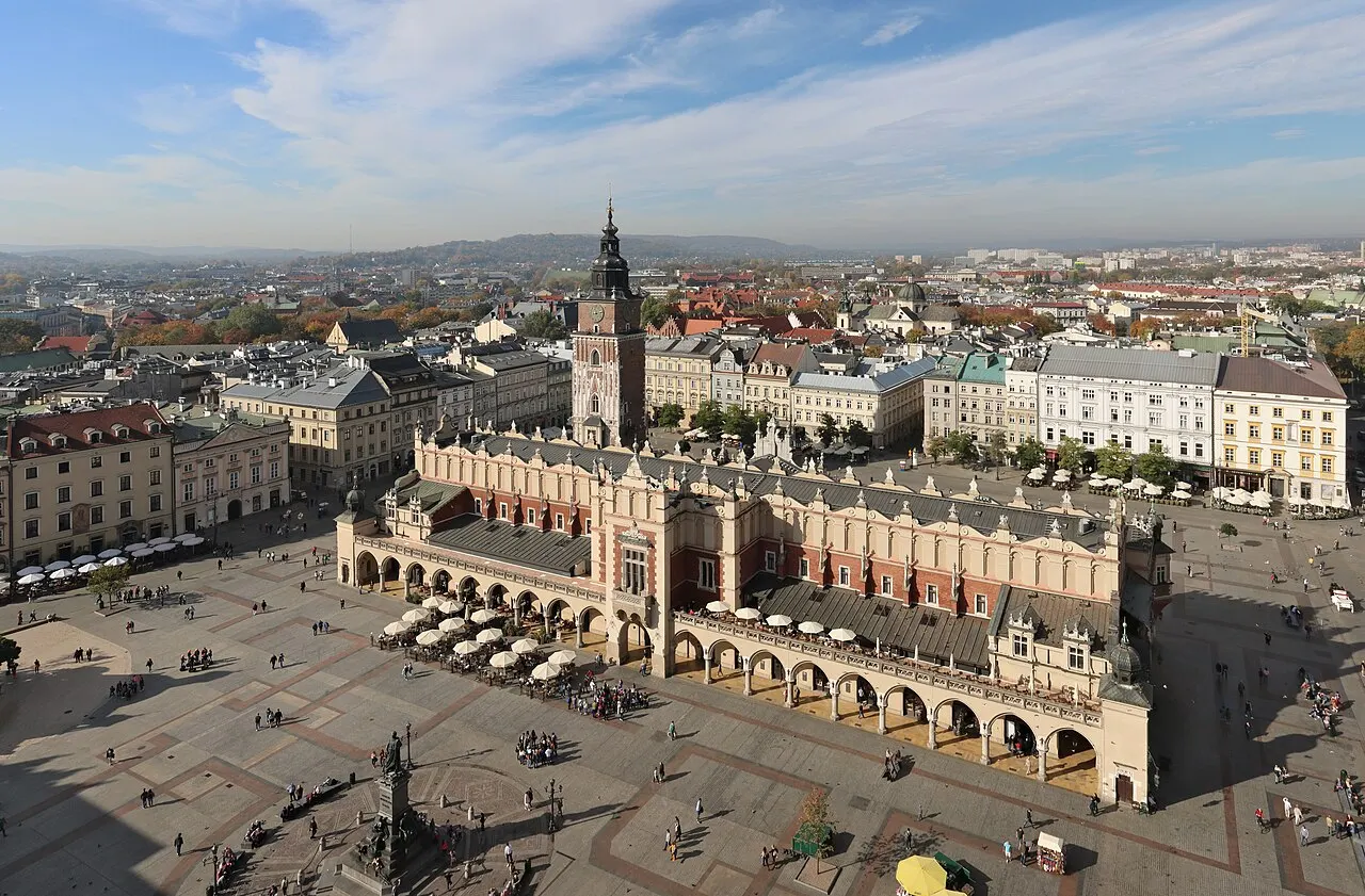 Vista aérea de uma grande praça de pedra cercada por prédios históricos coloridos, com um comprido salão comercial de arcadas ao centro, uma torre isolada ao lado e pessoas circulando entre mesas de cafés.