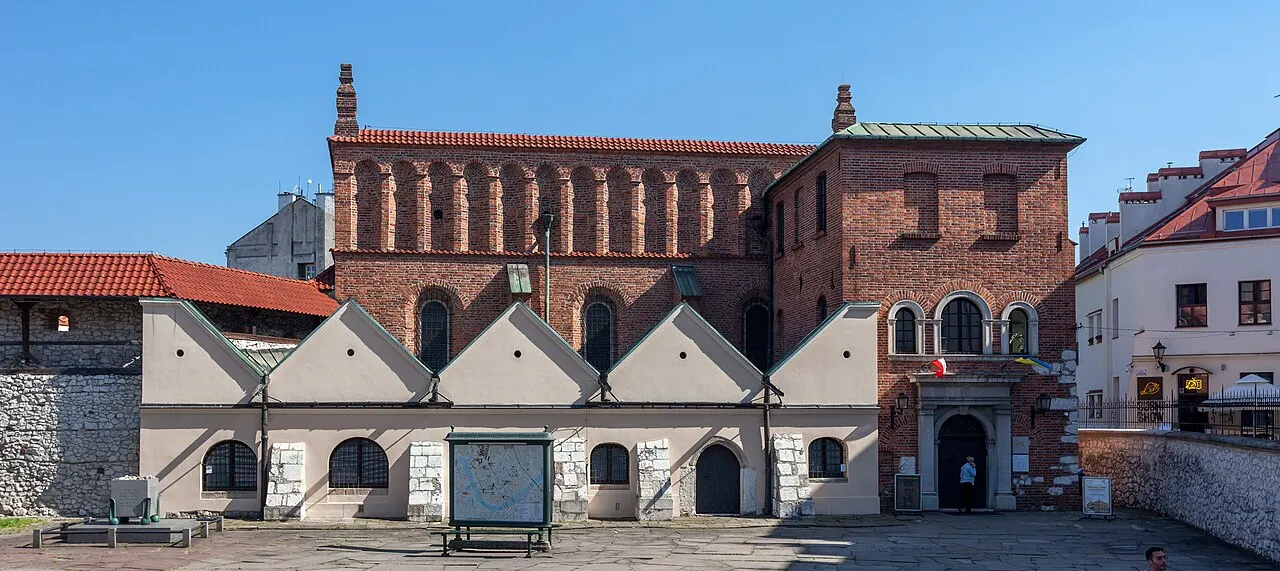 Prédio de tijolo vermelho da Sinagoga Velha de Kazimierz, com telhado de telha e uma fileira de arquinhos sob o beiral; na frente, um anexo baixo de fachadas claras e um trecho de muralha de pedra.