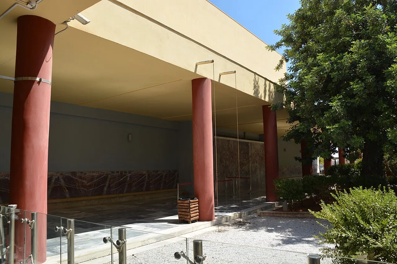 A entrada coberta do Museu Arqueológico de Heraklion, com colunas vermelhas cônicas, uma parede com relevo e árvores ao lado.