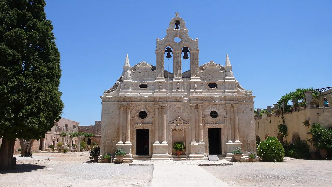 The pale stone facade of the Arkadi Monastery church, with columns, a three-bell gable and a cypress beside it.