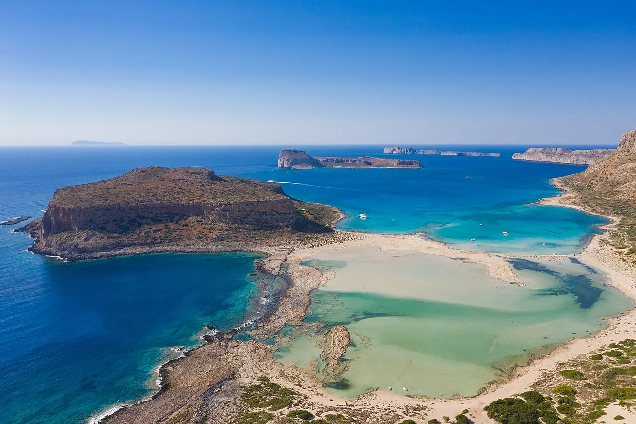 A lagoa de Balos vista do alto, com água turquesa rasa, faixas de areia branca e o cabo rochoso de Tigani.