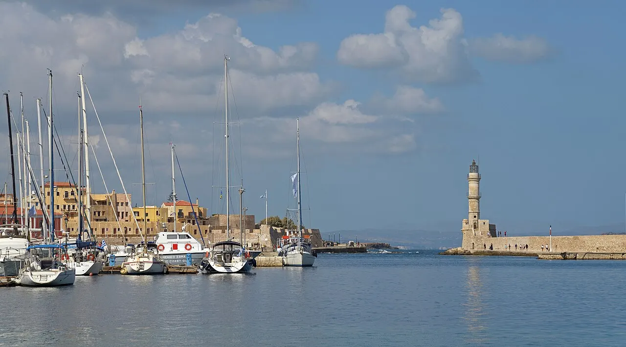 Chania's stone lighthouse at the end of the breakwater, with moored sailboats and the harbour houses on the left.