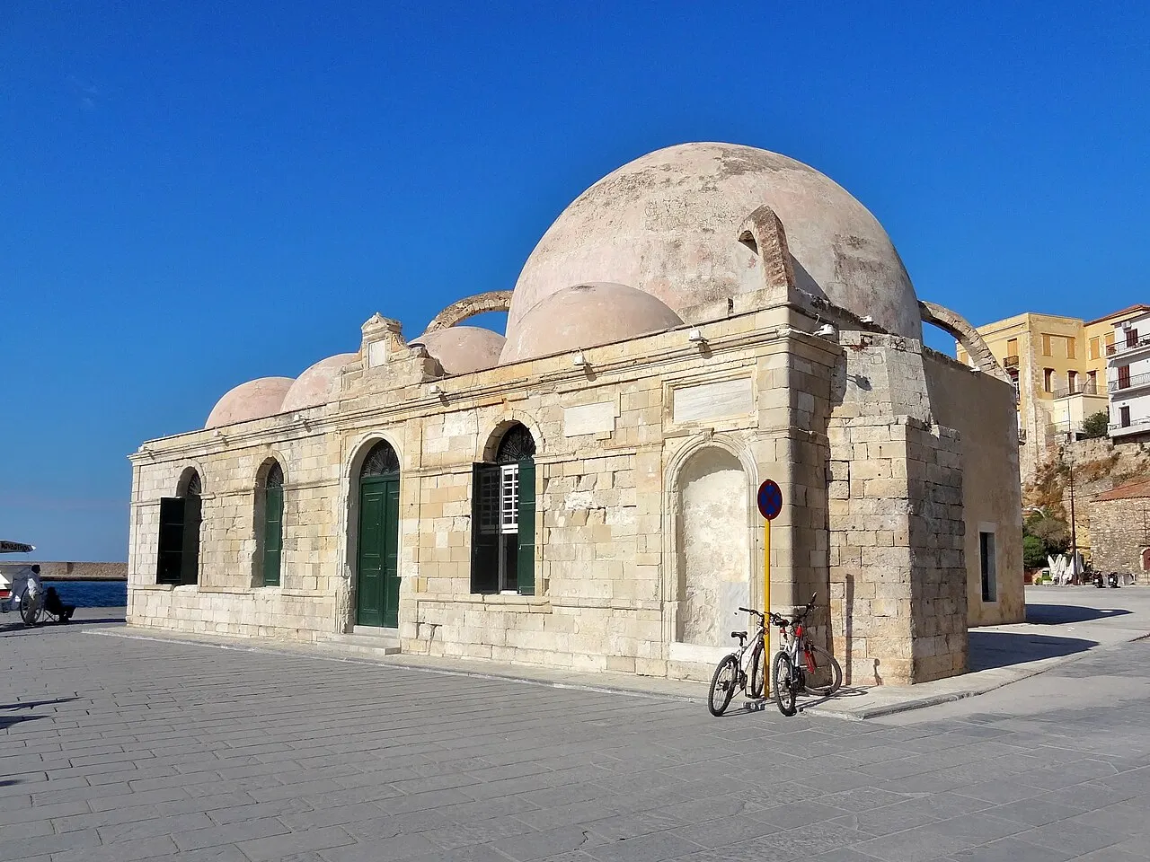 A mesquita de pedra clara com cúpula grande e pequenas cúpulas no porto de Chania, sob céu azul, com bicicletas na frente.