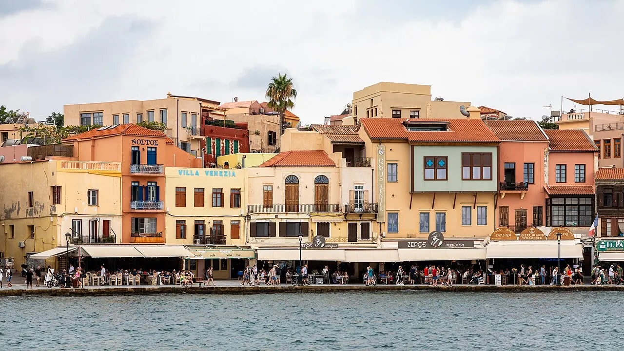 As casas coloridas da orla do porto veneziano de Chania, com toldos de restaurantes e pessoas no cais diante da água.
