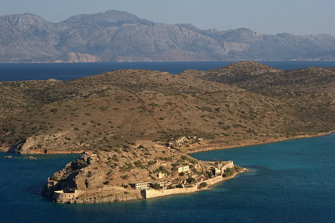 The small island of Spinalonga with its Venetian fortress, seen from above, in the blue sea before the peninsula and the mountains.