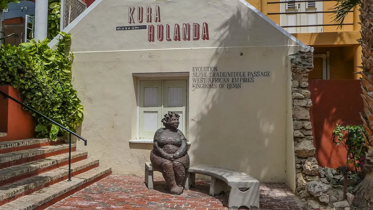 A entrada do Museum Kurá Hulanda, uma casa caiada com o nome na fachada e uma escultura de mulher sentada no banco em frente.