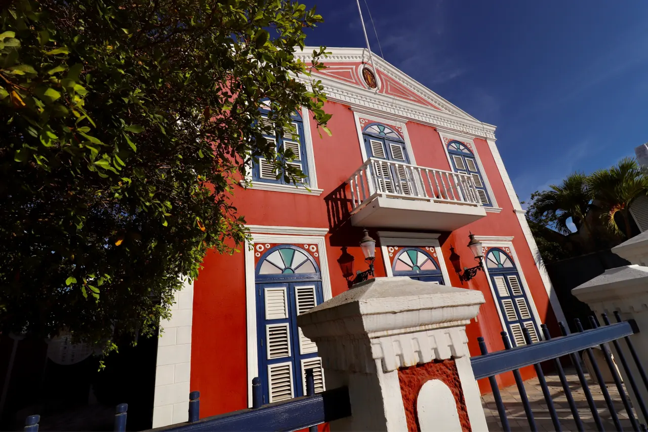 Uma casa antiga de Pietermaai pintada de vermelho com detalhes brancos, varanda e janelas altas, atrás de uma cerca e de árvores.