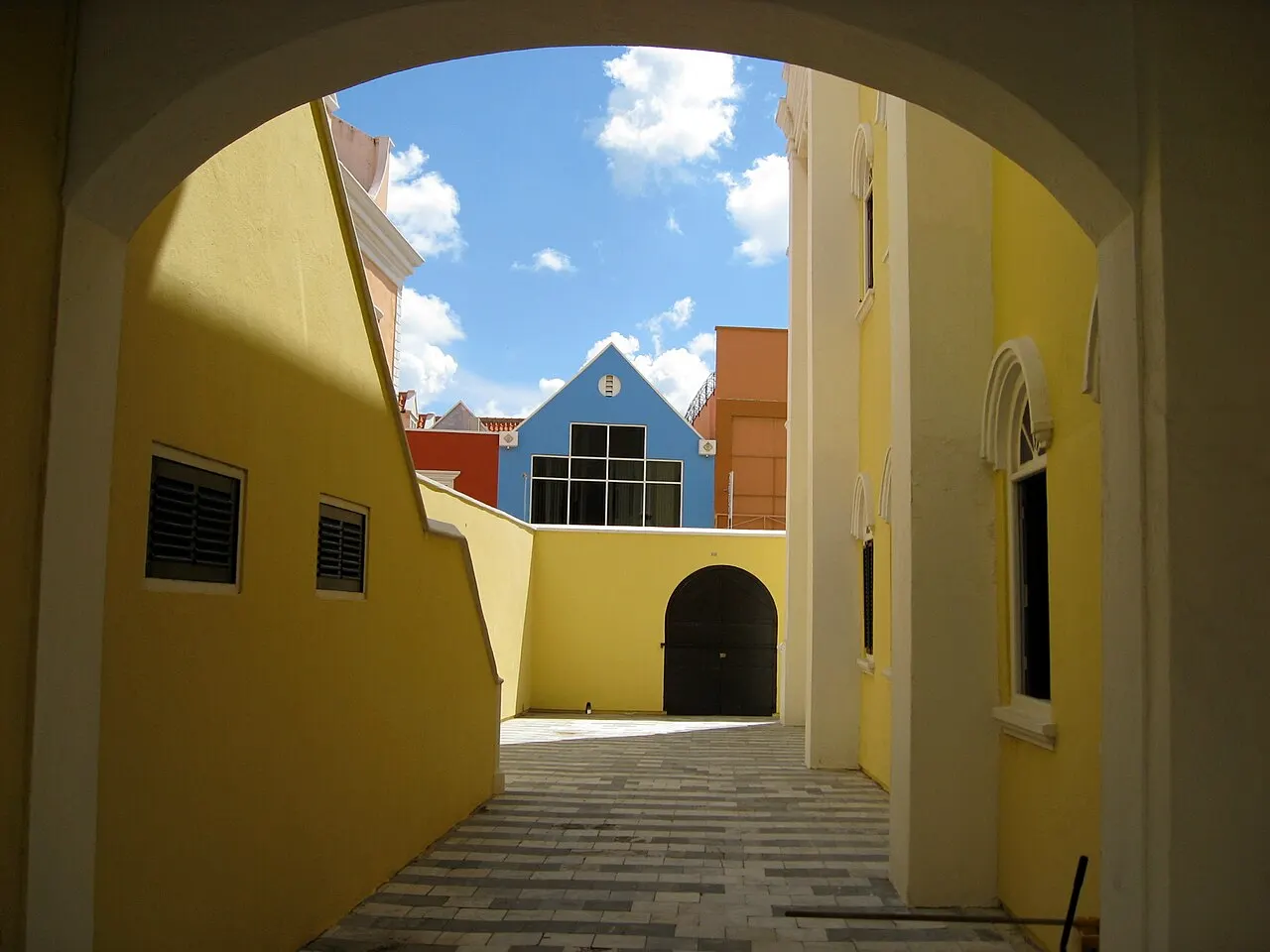 O pátio da sinagoga Mikvé Israel-Emanuel visto por um arco: paredes amarelas, janelas em arco e uma casa azul ao fundo.