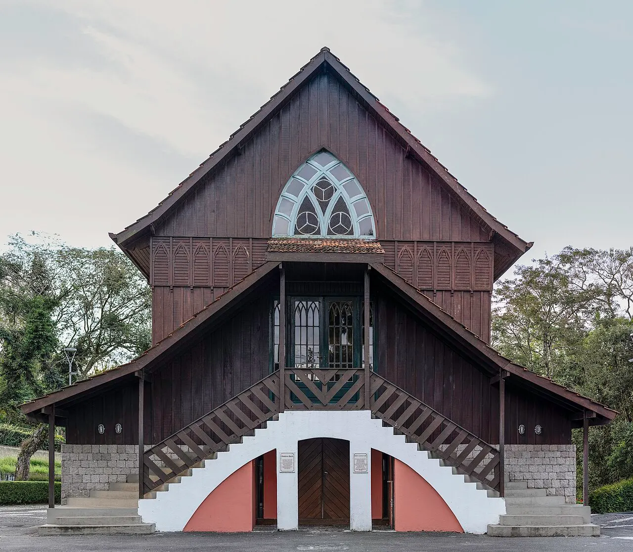 La capilla de madera oscura del Bosque Alemán, con techo puntiagudo y escalera de entrada, entre los árboles.