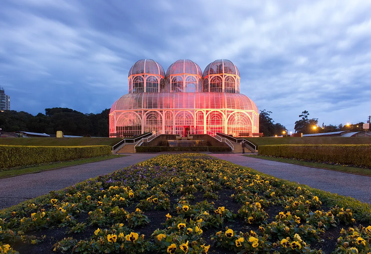 A estufa de vidro e metal do Jardim Botânico iluminada ao anoitecer, com os canteiros de flores amarelas na frente.