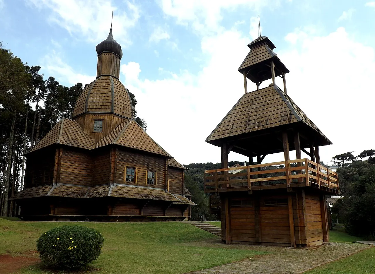 A igreja de madeira com cúpula em bulbo e o campanário do Memorial Ucraniano sobre o gramado, entre araucárias.