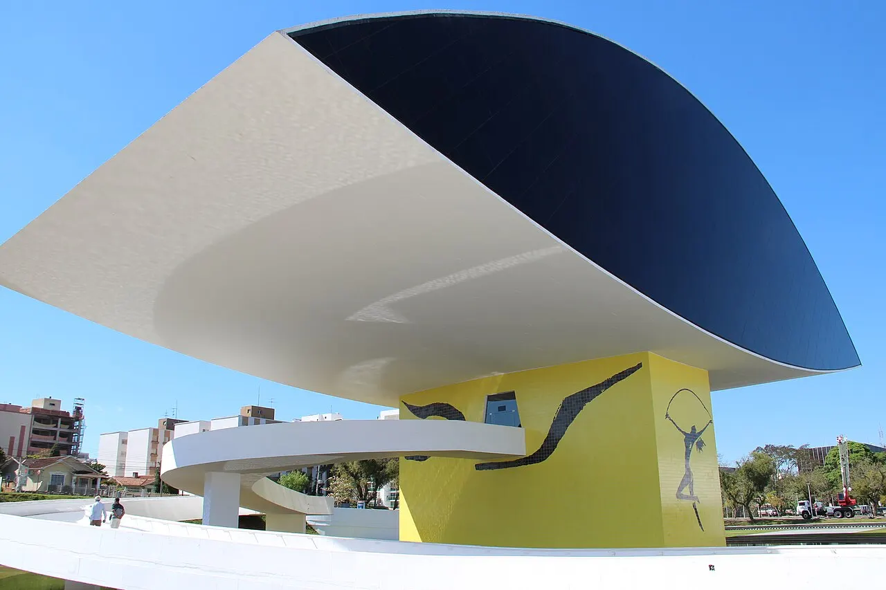O volume em forma de olho do Museu Oscar Niemeyer, branco e de vidro escuro, sobre o pilar amarelo pintado com figuras, sob céu azul.