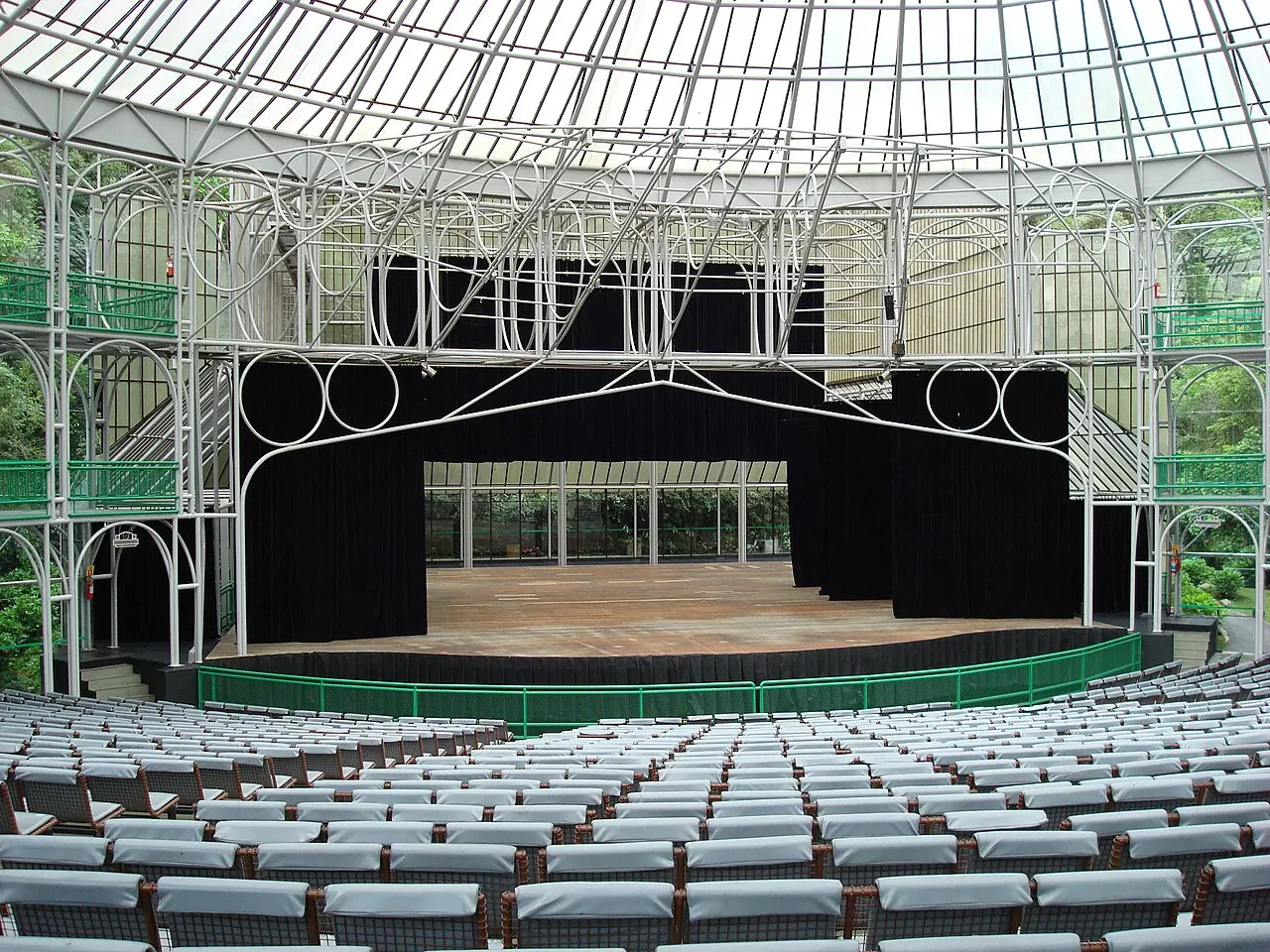 O palco e a plateia da Ópera de Arame sob a estrutura de tubos de metal e cobertura transparente, cercada de mata.