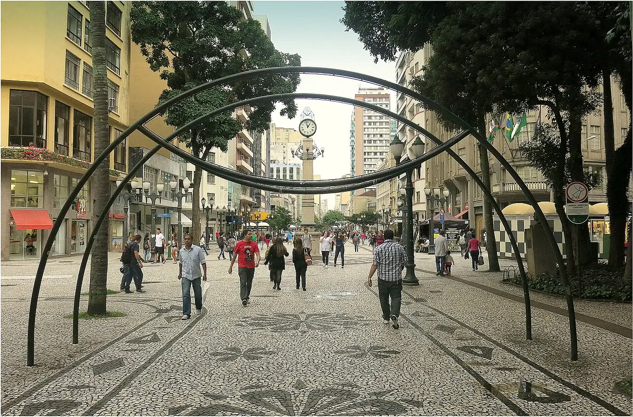 O calçadão de pedra portuguesa da Rua XV de Novembro, com arcos de metal sobre o caminho, árvores e gente passando.