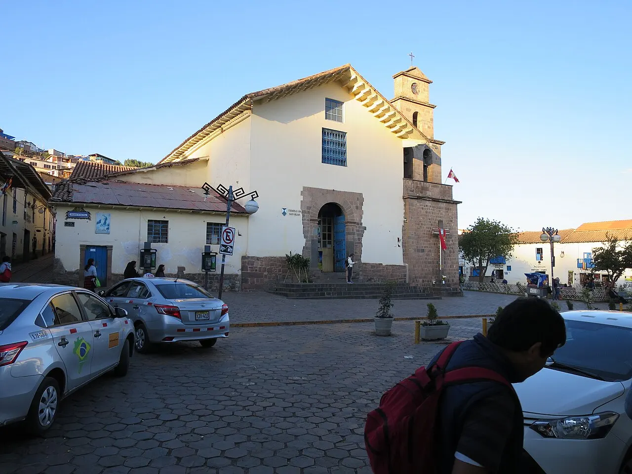 A igreja branca de San Blas, com a porta de pedra e o sino na empena, fica diante da praça e de carros estacionados.