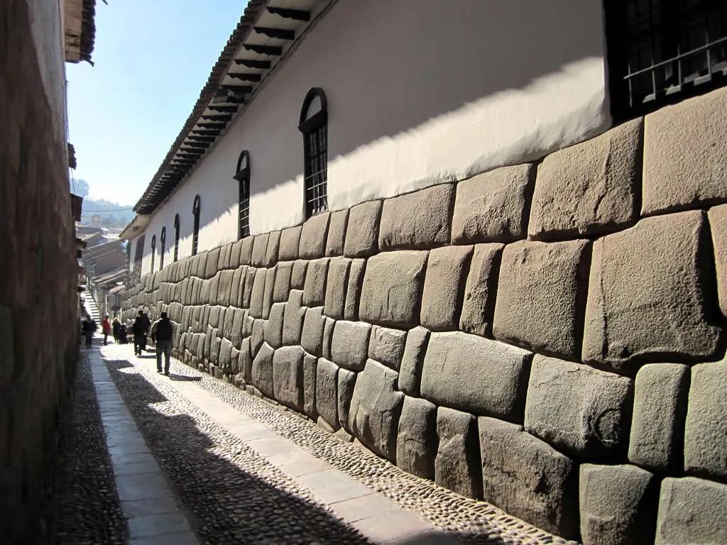 A Calle Hatunrumiyoc corre junto ao muro inca de pedras poligonais, sob a parede colonial branca com janelas gradeadas.