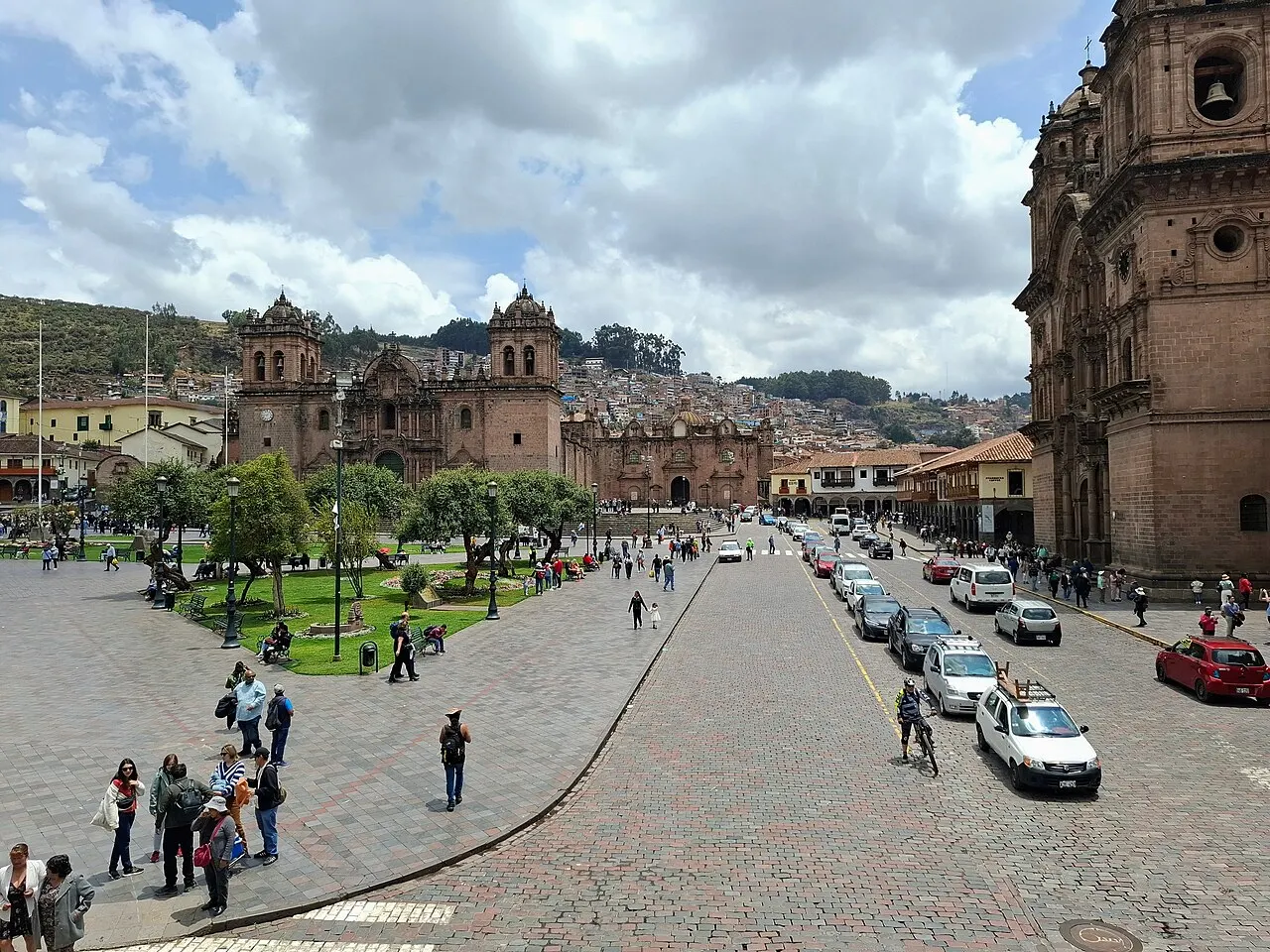 A catedral do Cusco em pedra escura, com duas torres de sino, fechando o lado norte da Plaza de Armas, a igreja da Companhia de Jesus à direita, o calçamento de pedra em primeiro plano e as casas subindo o morro ao fundo.