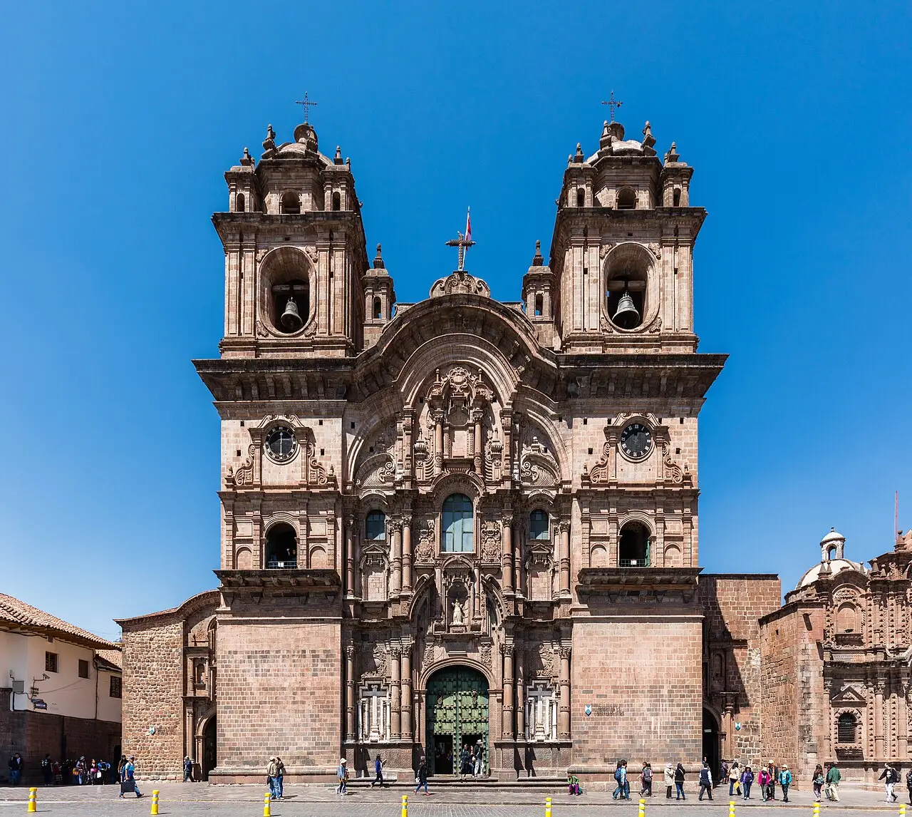 Fachada da igreja com duas torres de sinos, portal ao centro e pessoas na calçada, sob céu azul.