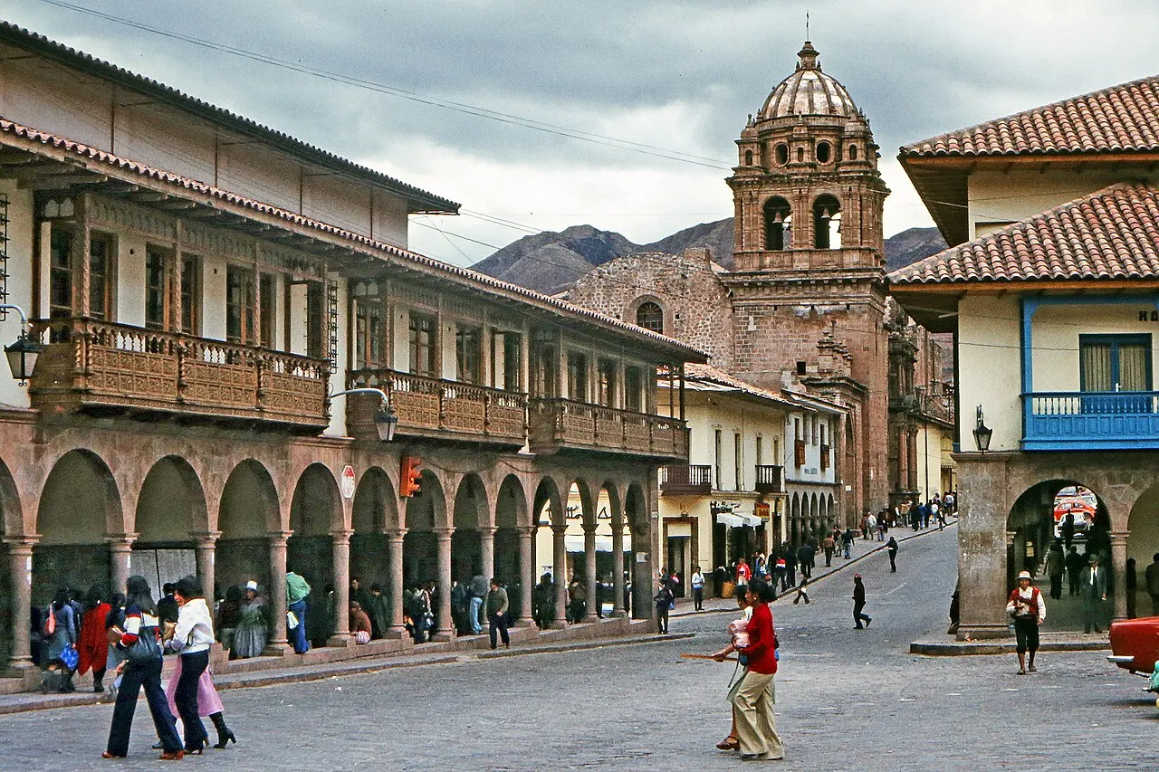Arcadas de pedra e sacadas de madeira em torno da Plaza de Armas de Cusco, com a torre de sino barroca ao fundo e a serra por cima dos telhados.