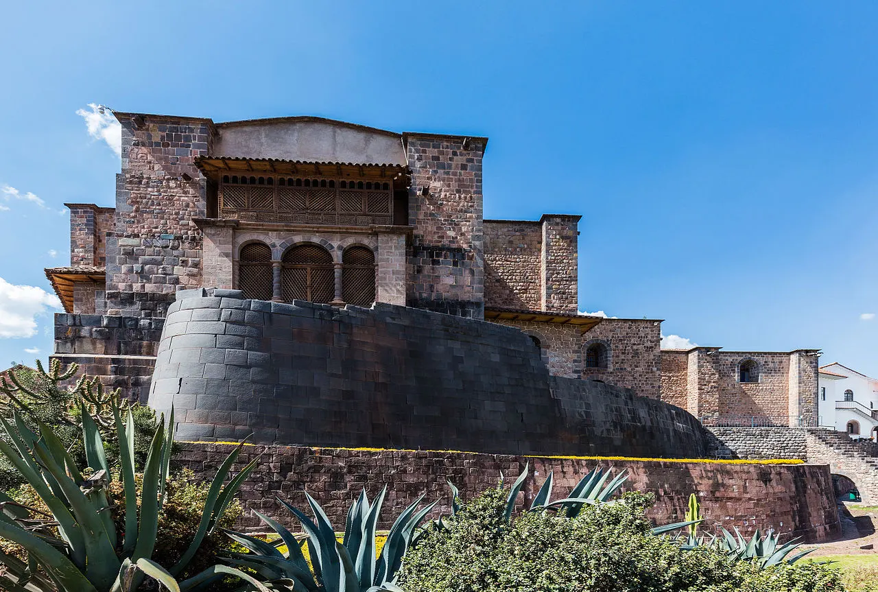 Um muro curvo de pedra escura com encaixes precisos na base, sustentando uma construção colonial de pedra clara com janelas em arco e uma varanda de madeira, plantas de agave em primeiro plano, sob céu azul.