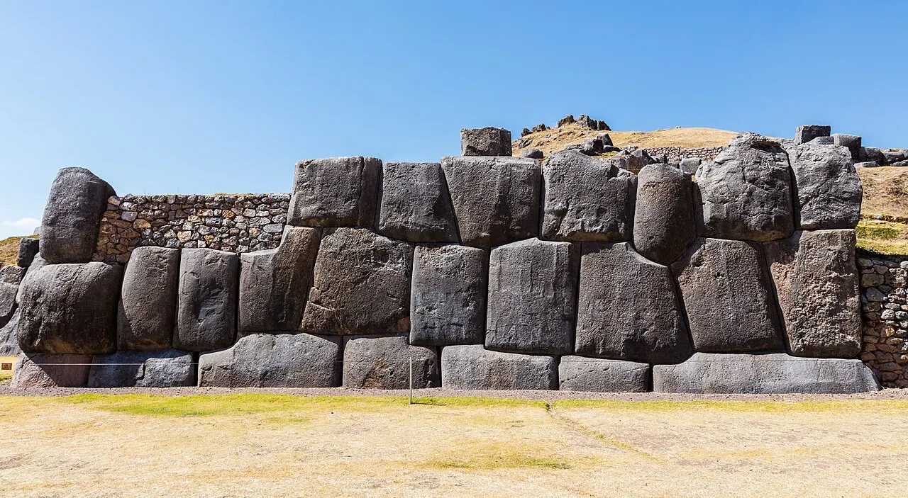 Uma muralha formada por enormes blocos de pedra de formas irregulares, encaixados uns nos outros sem argamassa, com um gramado seco em primeiro plano e uma colina ao fundo, sob céu azul limpo.
