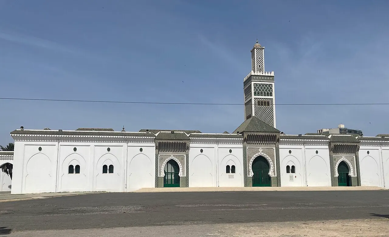 Muro branco comprido da Grande Mesquita de Dakar com portais verdes em arco recortado, faixas de mosaico verde e telhado de telha esmaltada, e o minarete alto vazado em filigrana à direita, sob céu azul.