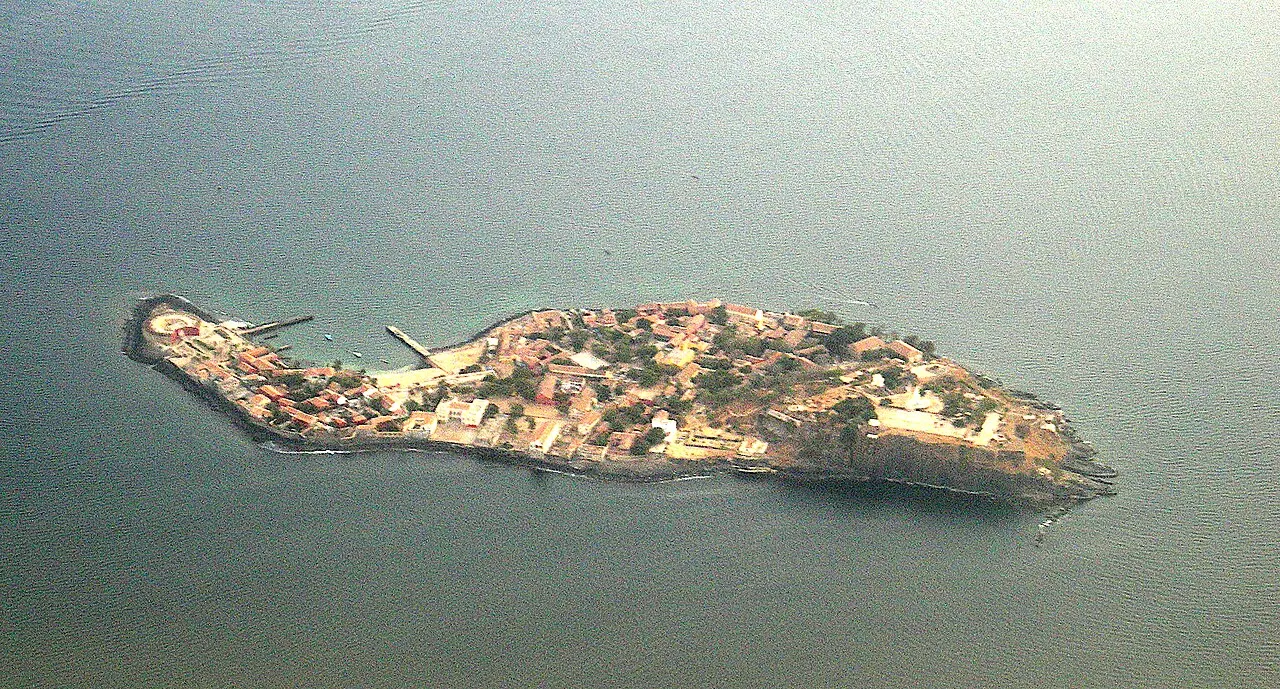 Vista aérea da ilha de Gorée, cercada pelo mar, com um forte arredondado à esquerda, o porto e os telhados do casario.