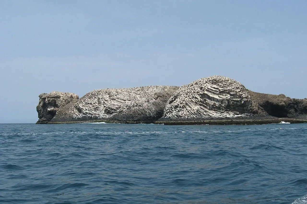 Ilha de rocha estratificada, branca e escura, cercada de mar azul, sem pessoas.