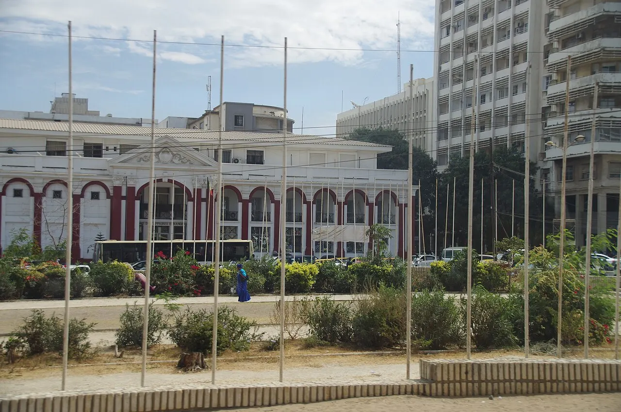 A Place de l'Indépendance junta uma fileira de mastros, o prédio de arcos vermelhos e brancos e um edifício branco alto ao lado.