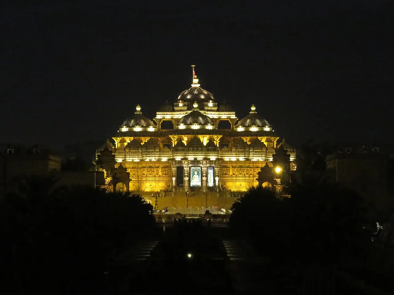 Templo de Akshardham iluminado à noite, com a cúpula central e palmeiras em silhueta na frente.
