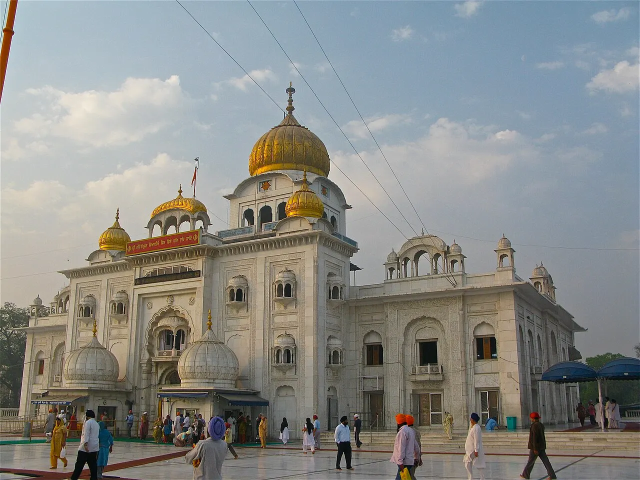 O Gurudwara Bangla Sahib tem cúpulas douradas sobre o mármore branco, e pessoas atravessam o pátio.