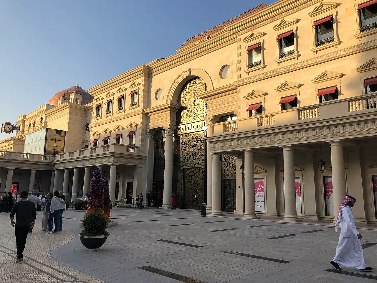 Praça pavimentada de Katara com arcadas de colunas e fachada de pedra clara acesa pelo sol baixo, um portal vazado dourado ao centro e pessoas atravessando.