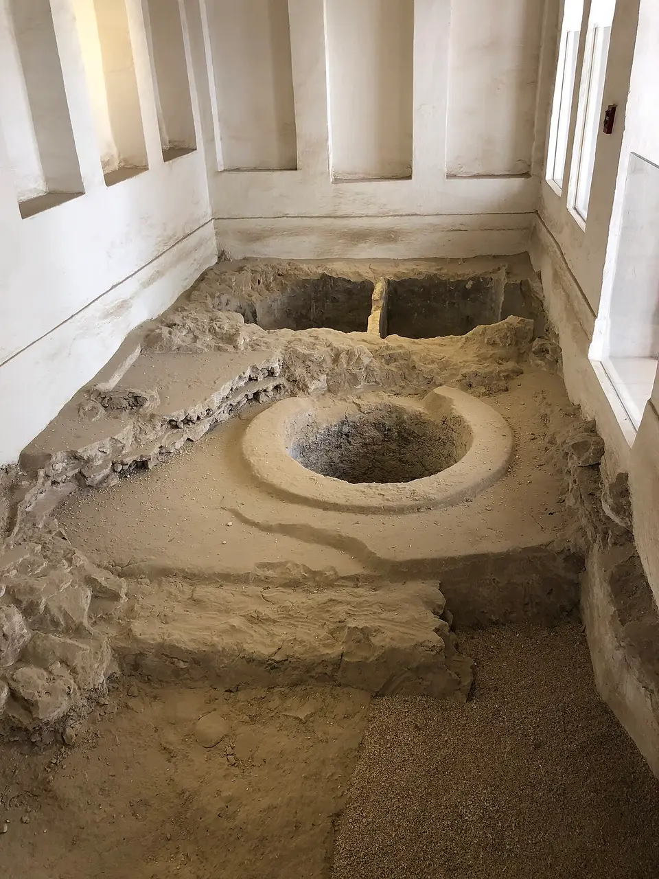 Restos de estruturas de terra, incluindo uma cavidade circular, preservados dentro de uma sala de paredes brancas.