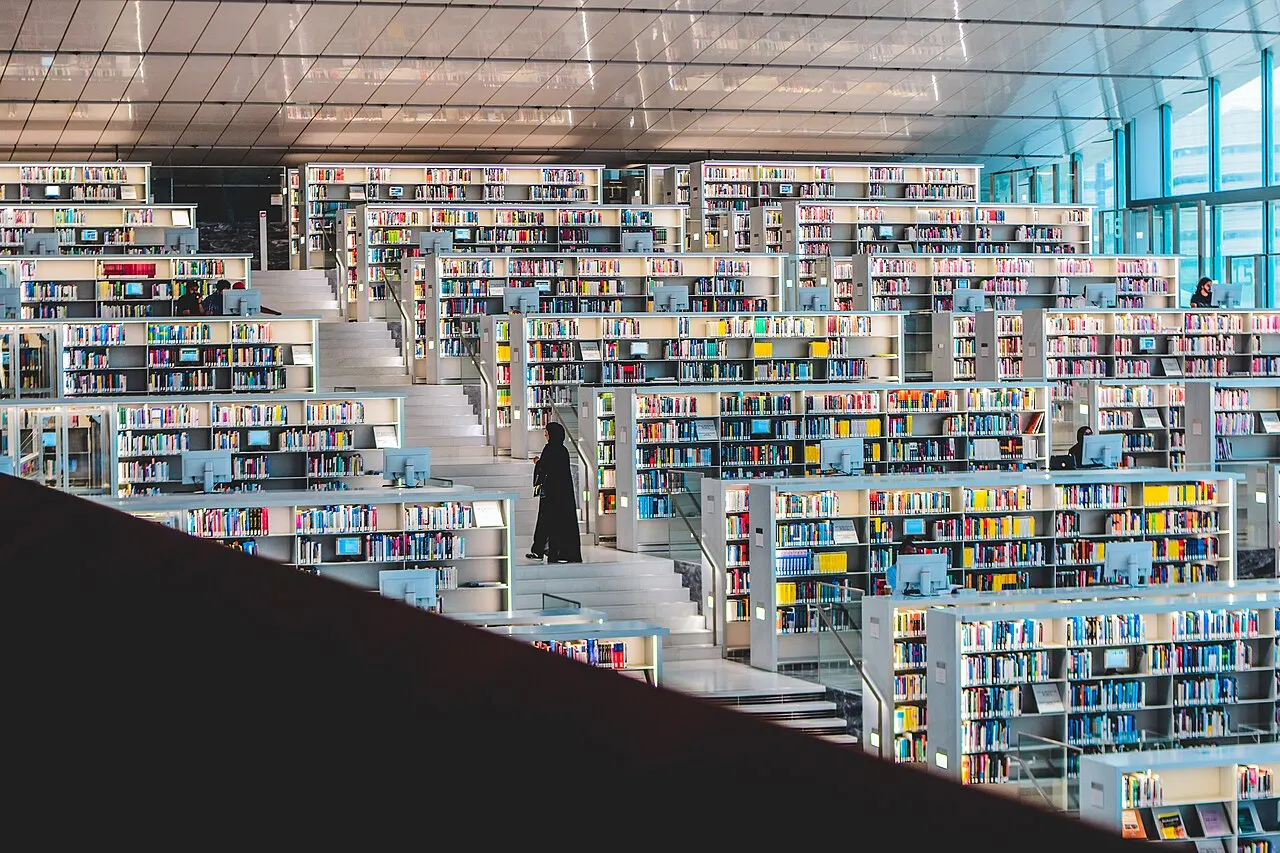 Estantes brancas em degraus, cheias de livros, ficam sob um teto de losangos, com uma pessoa de roupa escura no corredor e uma parede de vidro à direita.
