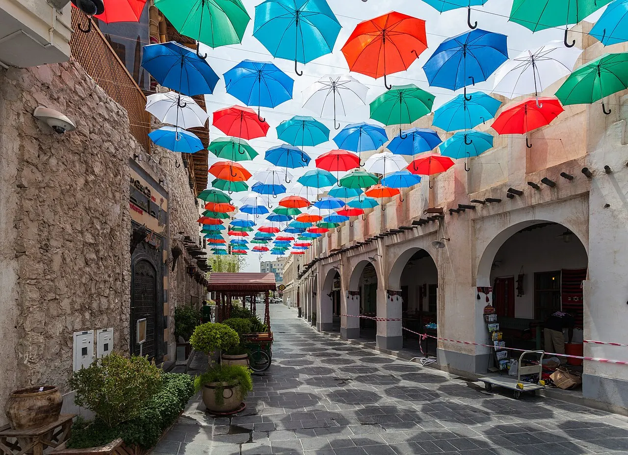 Uma rua estreita de pedra, com arcadas nas fachadas e dezenas de guarda-chuvas coloridos suspensos formando um teto acima do caminho, vasos de plantas e um carrinho na calçada, sob luz do dia.