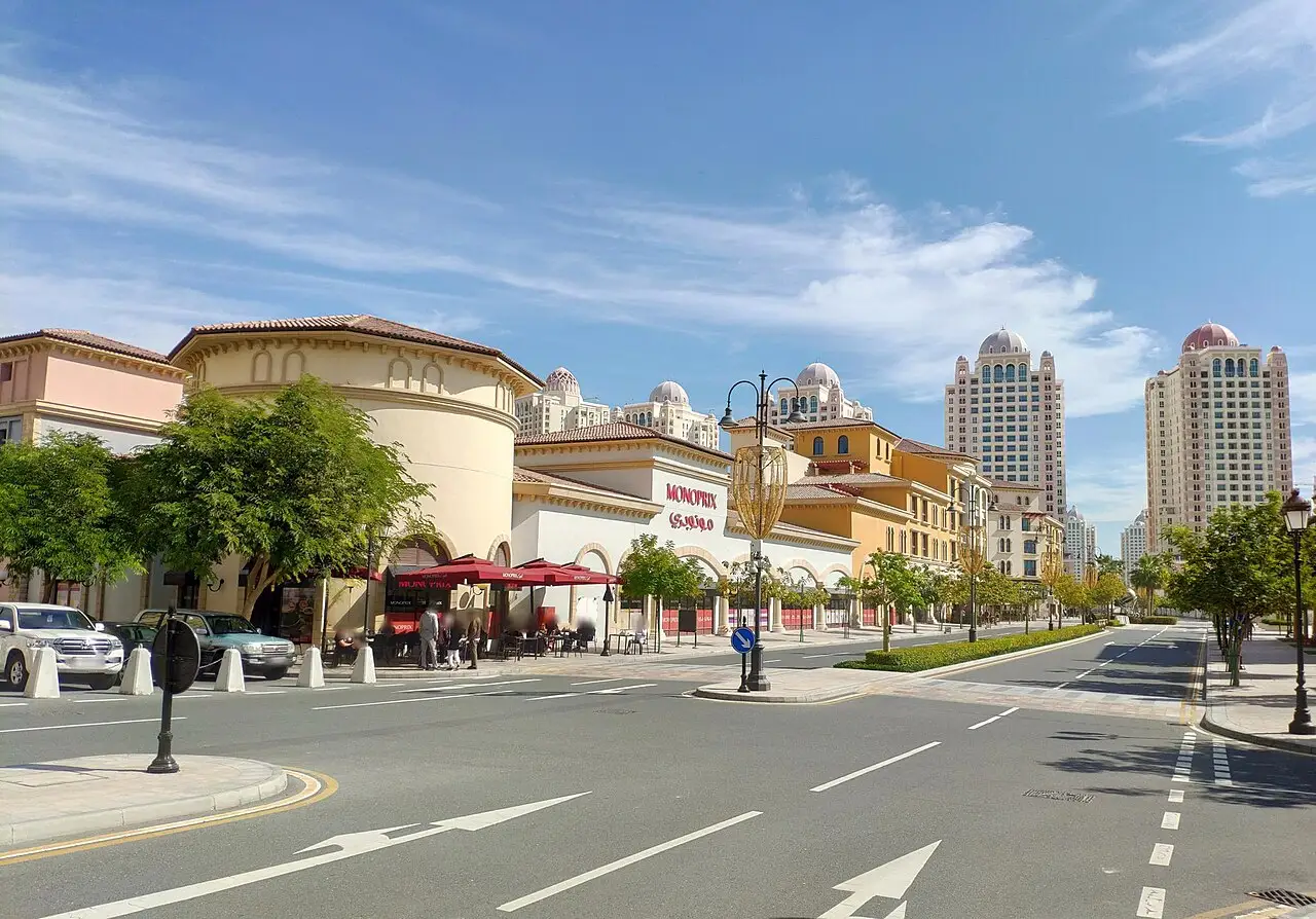 Avenida da The Pearl, com um edifício bege de canto arredondado, a loja Monoprix e torres claras ao fundo.