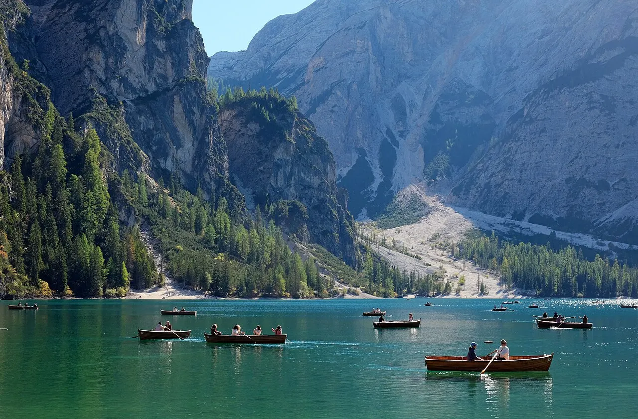 O Lago di Braies, verde-esmeralda, com barcos a remo de madeira e o paredão de rocha e floresta atrás.