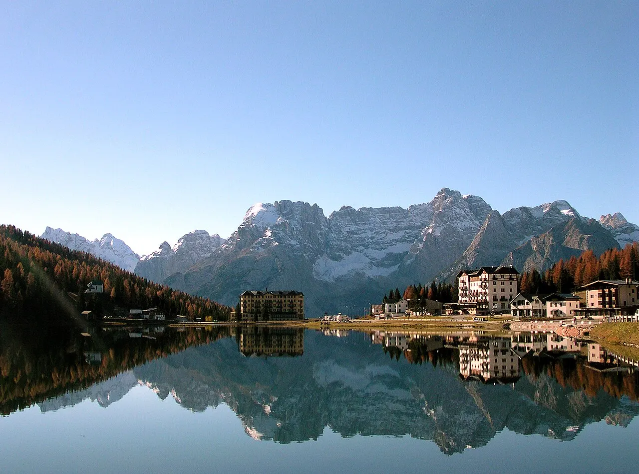 O Lago di Misurina com a água parada refletindo as montanhas e os hotéis da margem, entre lariços de outono.