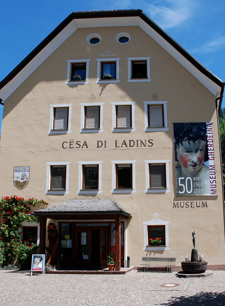 A fachada da Cësa di Ladins em Ortisei, com o nome pintado, janelas brancas e um cartaz do Museum Gherdëina.