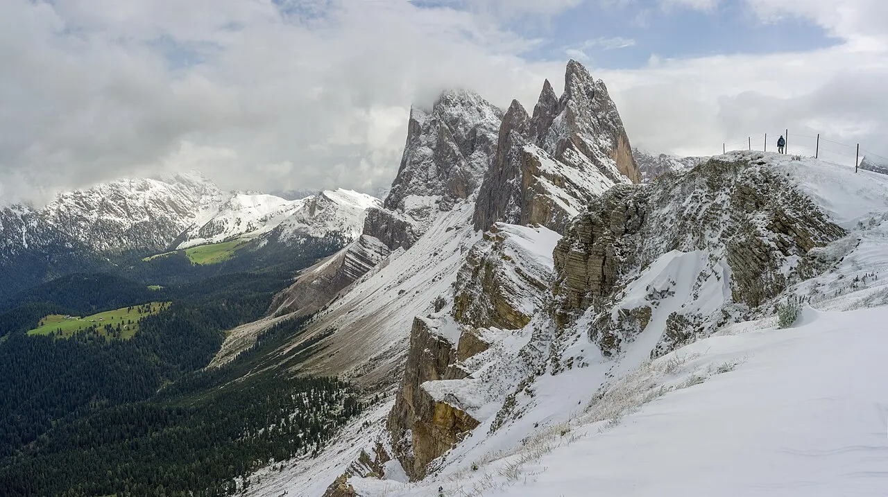 As agulhas de rocha das Odle vistas do Seceda, com a crista coberta de neve e uma pessoa pequena no alto do caminho.