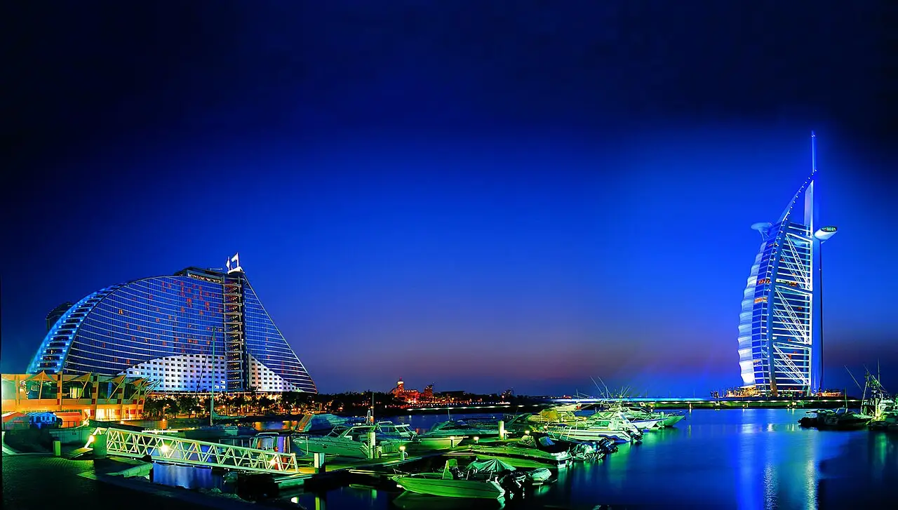 O Burj Al Arab iluminado à direita, um hotel em curva à esquerda e barcos no porto, sob céu azul escuro.