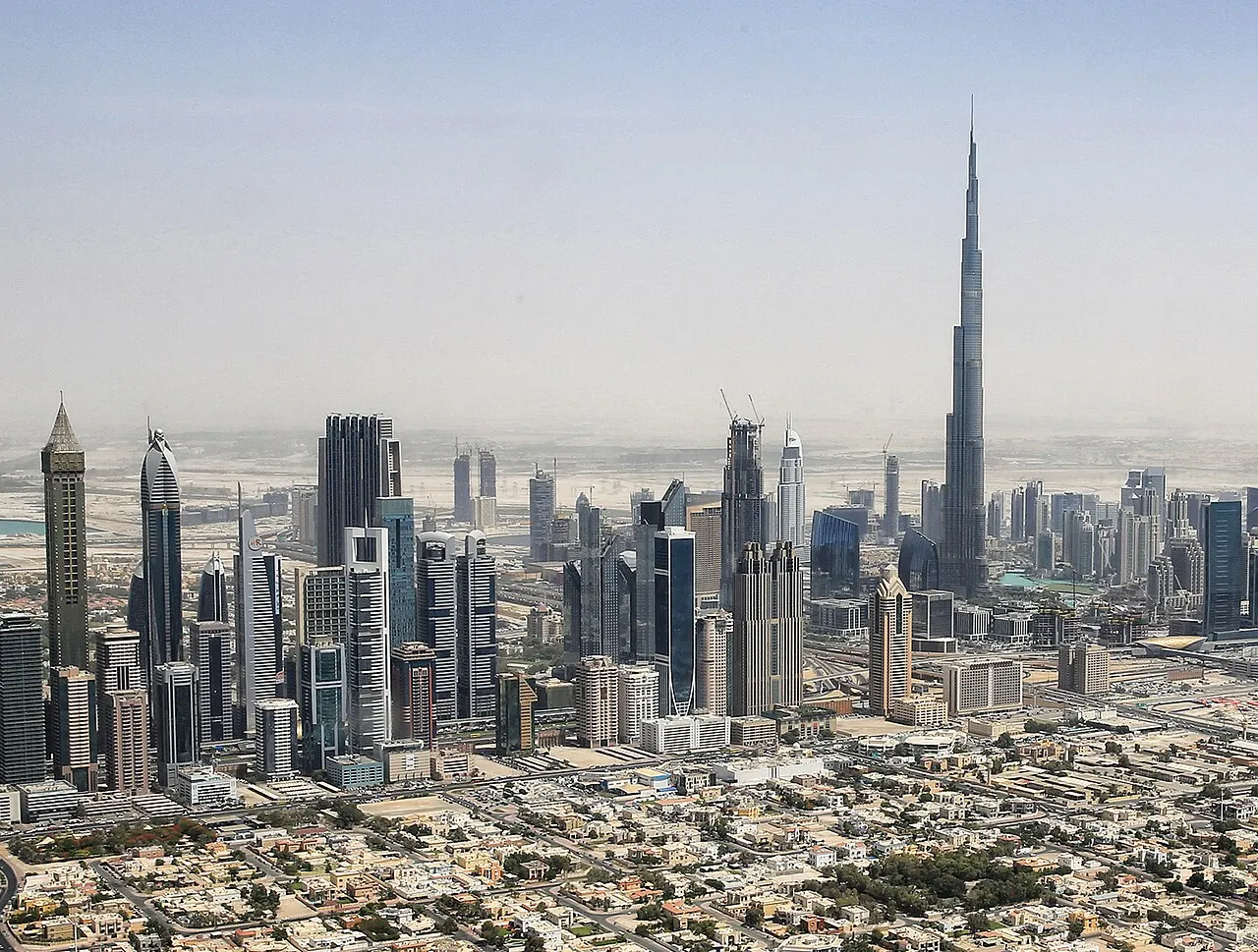 Vista aérea de uma cidade com dezenas de arranha-céus, com uma torre muito mais alta e afilada se destacando no centro da paisagem, deserto ao fundo sob névoa e céu claro.