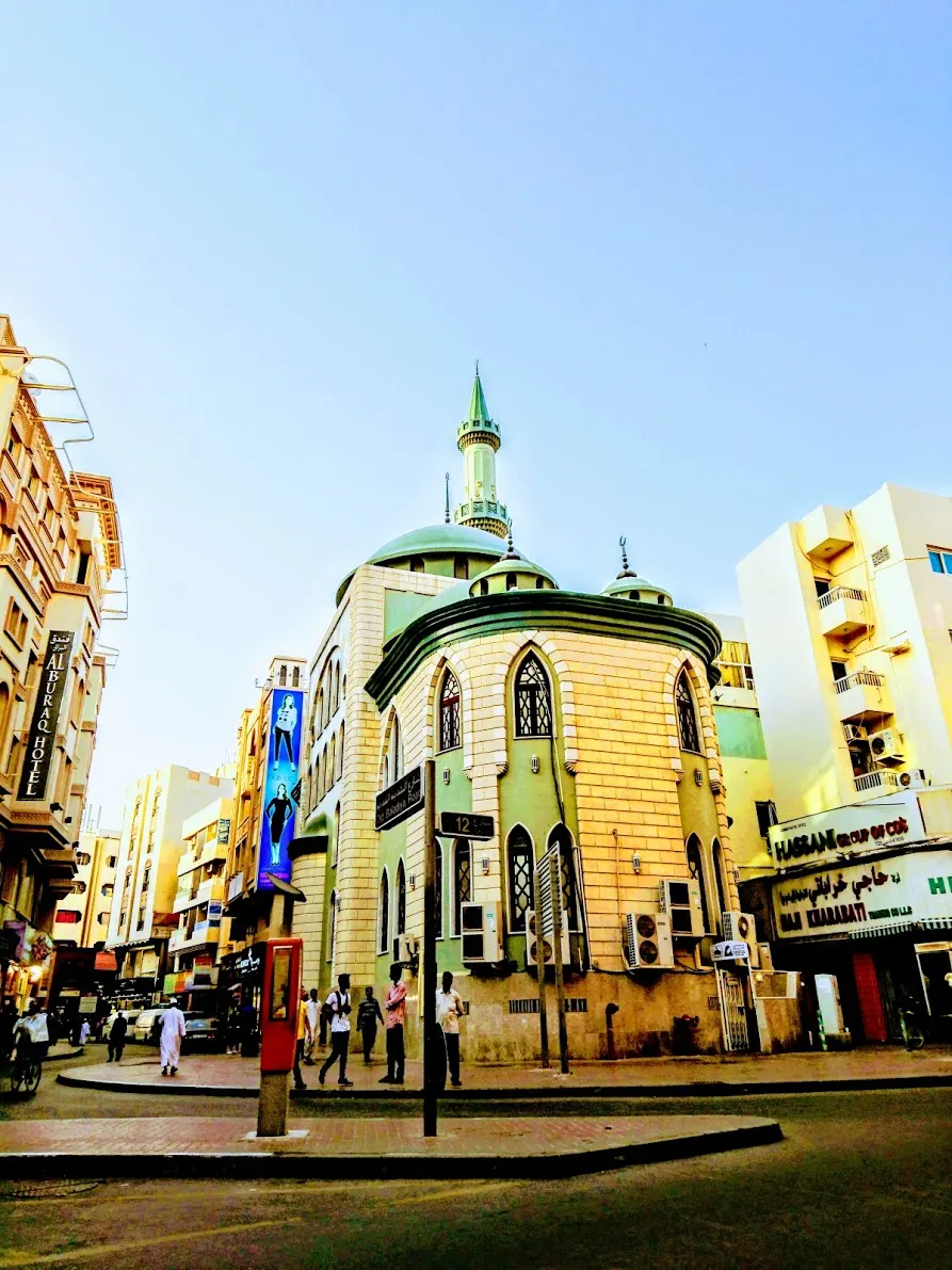 Prédio bege de esquina com uma torre octogonal e cúpula verde, com letreiros ao fundo.