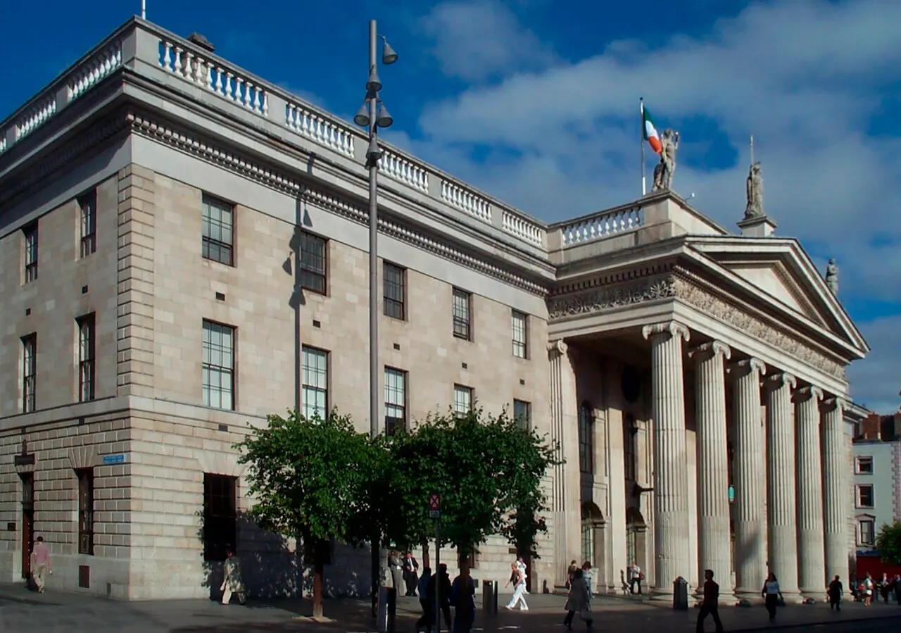 Fachada de granito do General Post Office de Dublin com pórtico de colunas jônicas, estátuas no frontão e a bandeira irlandesa hasteada, vista da esquina da O'Connell Street.