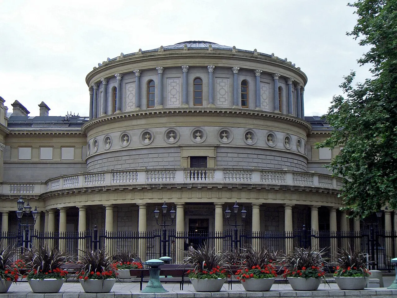 A fachada curva de um prédio neoclássico, com uma rotunda de colunas sob uma cúpula de vidro, um pórtico de entrada mais baixo à frente, grades de ferro e jardineiras com flores vermelhas, sob céu nublado.