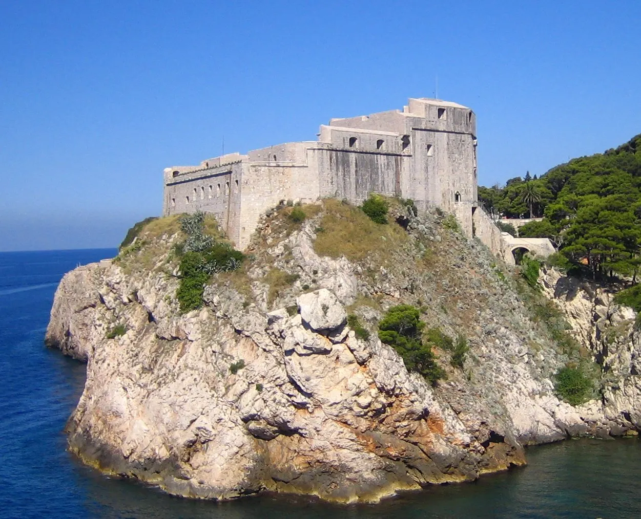 Um forte de pedra clara fica no alto de um rochedo, com o mar dos dois lados e pinheiros à direita, sob céu azul.