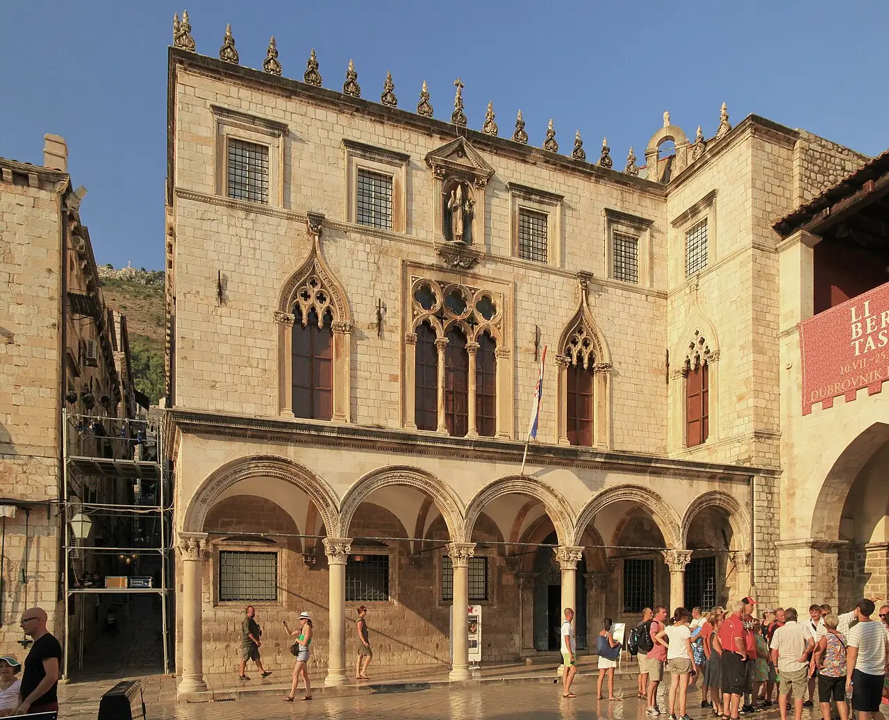 Fachada do Palácio Sponza, com arcada no térreo, janelas góticas e uma estátua no nicho, e pessoas na frente.