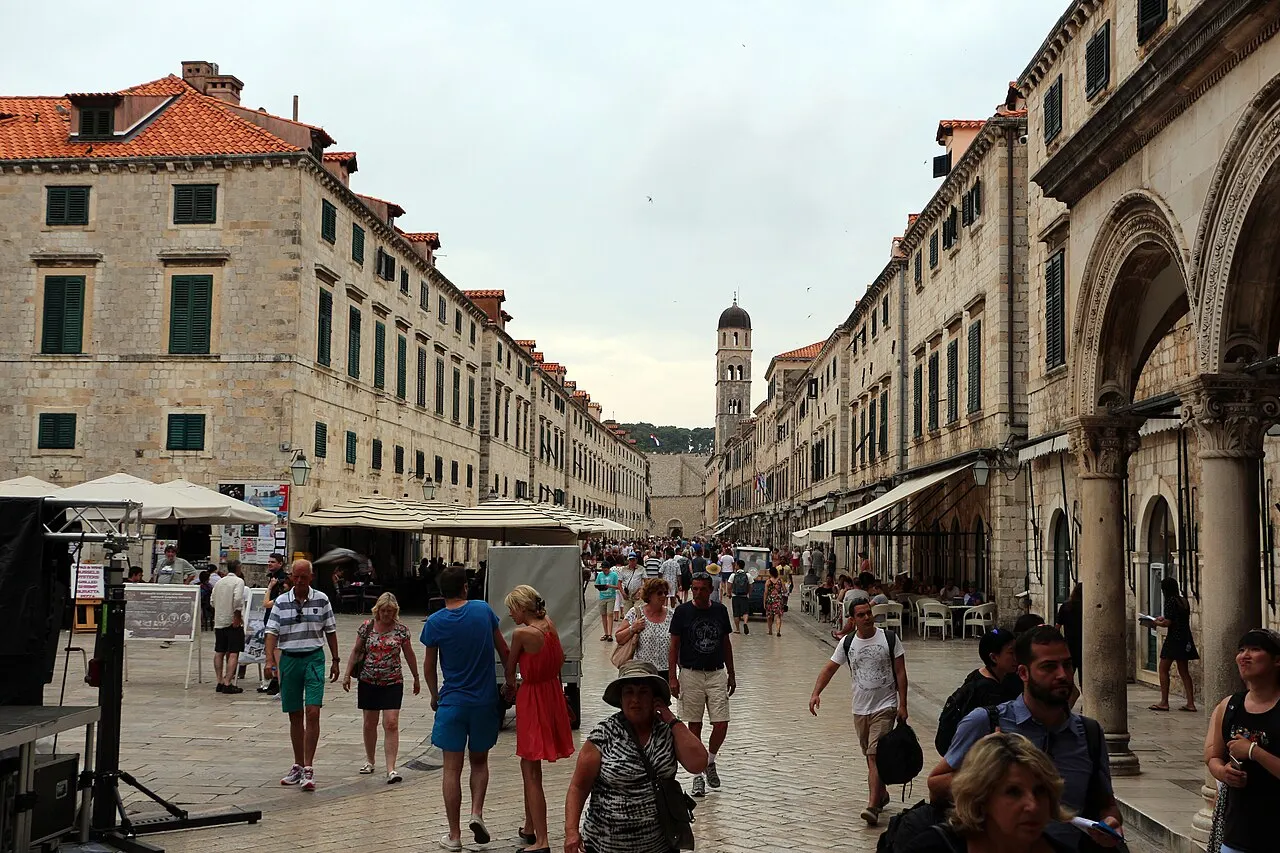 Uma rua larga de pedra polida, ladeada por fachadas uniformes de pedra clara com janelas de veneziana verde, mesas de café sob toldos e uma torre de sino ao fundo, com pessoas caminhando sob céu nublado.