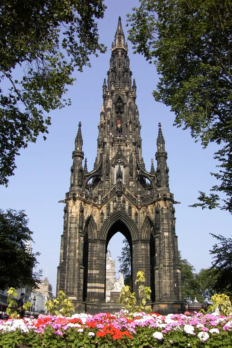 Torre gótica alta de pedra escurecida, com pináculos e uma estátua sob o arco central, e flores à frente.