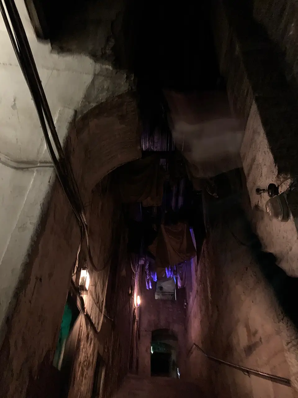 Um beco subterrâneo estreito, de paredes de pedra, com o chão escuro e uma luz roxa no fim.